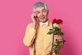 Handsome senior man talks via cellphone, wears yellow shirt with braces and bowtie, discusses date with friend, holds bouquet of