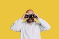 Handsome serious middle-aged man in a white shirt smiles and holds black binoculars on a yellow background with copy space Royalty Free Stock Photo