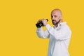 Handsome serious middle-aged man in a white shirt smiles and holds black binoculars on a yellow background with copy space Royalty Free Stock Photo