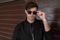 Handsome sexy young hipster man in trendy sunglasses in a black jacket with a stylish hairstyle with a clock poses near a wooden Royalty Free Stock Photo