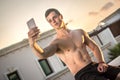 Handsome shirtless muscular young man taking selfie with smart phone while sitting on rooftop during sunset. Royalty Free Stock Photo