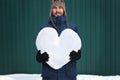 Handsome smiling bearded hipster man in winter clothes holding a big heart made of snow, on green wall background. Fall in love.