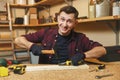 Handsome smiling young man working in carpentry workshop at wooden table place with piece of wood Royalty Free Stock Photo