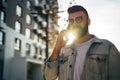 Handsome hipster businessman with beard, in denim jacket and trendy glasses walks around city and calls on mobile phone. Royalty Free Stock Photo