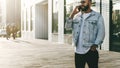 Handsome hipster businessman with beard, in denim jacket and trendy glasses walks around city and calls on mobile phone Royalty Free Stock Photo