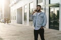 Handsome hipster businessman with beard, in denim jacket and trendy glasses walks around city and calls on mobile phone Royalty Free Stock Photo