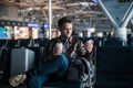 Handsome smiling man in casual wear holding luggage and messaging through his mobile phone while sitting at the hall of Royalty Free Stock Photo