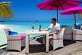 Handsome smiling man is sitting in beach bar on the Maldives sandy beach and holding colorful fruity drink. Luxury travel. Tourism