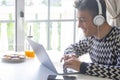 Handsome, smiling teenager using laptop at home in front of the window wearing headphones. Millenial generation boy enjoying