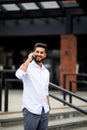 Handsome smiling young indian man dressed casually spending time outdoors at the city, using mobile phone while walking down Royalty Free Stock Photo