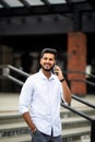 Handsome smiling young indian man dressed casually spending time outdoors at the city, using mobile phone while walking down Royalty Free Stock Photo