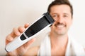 Handsome smiling young man with towel on shoulders showing electric shaving razor and looking at camera, close-up.