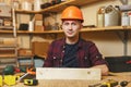 Handsome smiling young man working in carpentry workshop at wooden table place with piece of wood