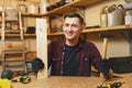 Handsome smiling young man working in carpentry workshop at wooden table place with piece of wood