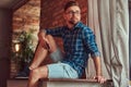 Handsome stylish hipster holds a smartphone while sitting on a table in a room with a loft interior.