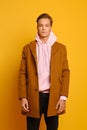 Handsome stylish man in brown trenchcoat and pink sweatshot with hood posing over yellow background