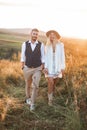 Handsome stylish man in shirt, vest and pants and pretty boho woman in dress, jacket and hat walking in the field with