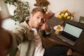 Handsome stylish young man takes selfie in cafe while working or studying remotely with laptop, shows call phone hand Royalty Free Stock Photo