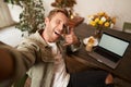 Handsome stylish young man takes selfie in cafe while working or studying remotely with laptop, shows call phone hand Royalty Free Stock Photo