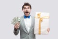 Handsome suprised young bearded man in gray suit and blue bow tie standing and holding gift box with yellow bow and many cash