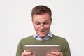 Handsome surprised young man wearing green sweater looking on tablet