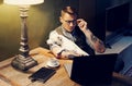 Handsome tattooed man in eyeglasses working at home on laptop while sitting at the wooden table with cute dog on his