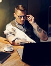 Handsome tattooed man in eyeglasses working at home on laptop while sitting at the wooden table with cute dog on his Royalty Free Stock Photo