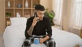 Handsome, tattooed young adult latin man comfortably relaxing on bed, sipping java, seriously concentrating on phone talk Royalty Free Stock Photo