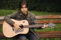 Handsome teenage playing acoustic guitar outdoor in Autumn time. Boy sitting on bench and playing music