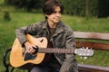 Handsome teenage playing acoustic guitar outdoor in Autumn time. Boy sitting on bench and playing music