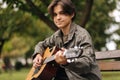 Handsome teenage playing acoustic guitar outdoor in Autumn time. Boy sitting on bench and playing music