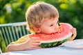 Handsome toddler boy eating fresh red watermelon. Yummy portrait of smiling child sits by the table in garden. Healthy Royalty Free Stock Photo