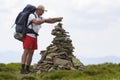 Handsome tourist man with backpack making pyramid pile of stones on lit by summer sun green grassy mountain valley on light blue c