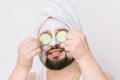 Handsome unshaven man with white towel on his head and clay mask on skin, covers eyes with two slices of cucumber Royalty Free Stock Photo