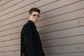 Handsome urban young man model in youth casual black denim jacket in stylish glasses with trendy hairstyle stands near wooden wall Royalty Free Stock Photo