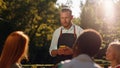 A handsome waiter with his hair tied back is taking an order from a table that s outside, with beautiful trees in the Royalty Free Stock Photo