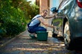 Handsome young adult man in blue coverall washing his car with foamy detergent and sponge rag, smiling looking at camera Royalty Free Stock Photo