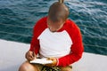 Handsome young African-American hipster man relaxing on the beach and making text notes in paper notebook. Blue sea on Royalty Free Stock Photo