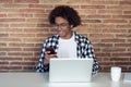 Handsome young afro-american man with eyeglasses using his smartphone while working with laptop at home Royalty Free Stock Photo