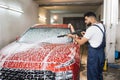 Handsome young bearded man worker, wearing protective clothes and gloves, washing modern red car Royalty Free Stock Photo