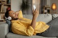 Handsome young black guy in wireless headphones listening to music online on smartphone, lying on couch at home Royalty Free Stock Photo