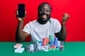 Handsome young black man playing poker holding smartphone screaming proud, celebrating victory and success very excited with Royalty Free Stock Photo