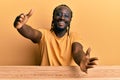 Handsome young black man wearing casual clothes sitting on the table looking at the camera smiling with open arms for hug Royalty Free Stock Photo