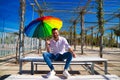 Handsome young blue-eyed gay man is sitting in a park and holding a rainbow-coloured umbrella in his hand. The man is happy.