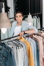 Young boutique owner leaning at hangers with clothes and smiling at camera