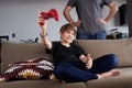 Handsome young boy playing video game console seated on a sofa while his father standing close in living room at home Royalty Free Stock Photo