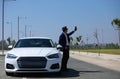 Handsome young businessman with beard and blue suit standing next to his white sports car. The man wears sunglasses and takes Royalty Free Stock Photo