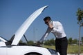 Handsome young businessman with a beard looks at the engine of his car which is broken down on the road and phones the insurance