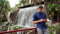 Handsome young businessman outdoors, sliding smartphone touchscreen, waterfall