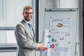 handsome young businessman in suit and eyeglasses pointing at whiteboard with charts and smiling Royalty Free Stock Photo